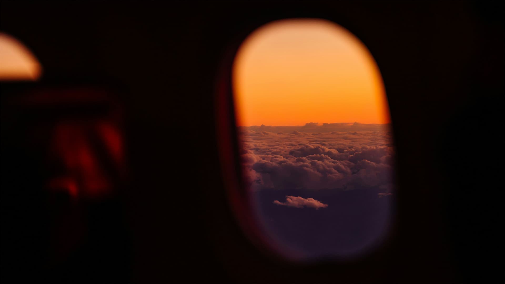 View through an airplane window over a sea of clouds at sunset, with an orange sky on the horizon.